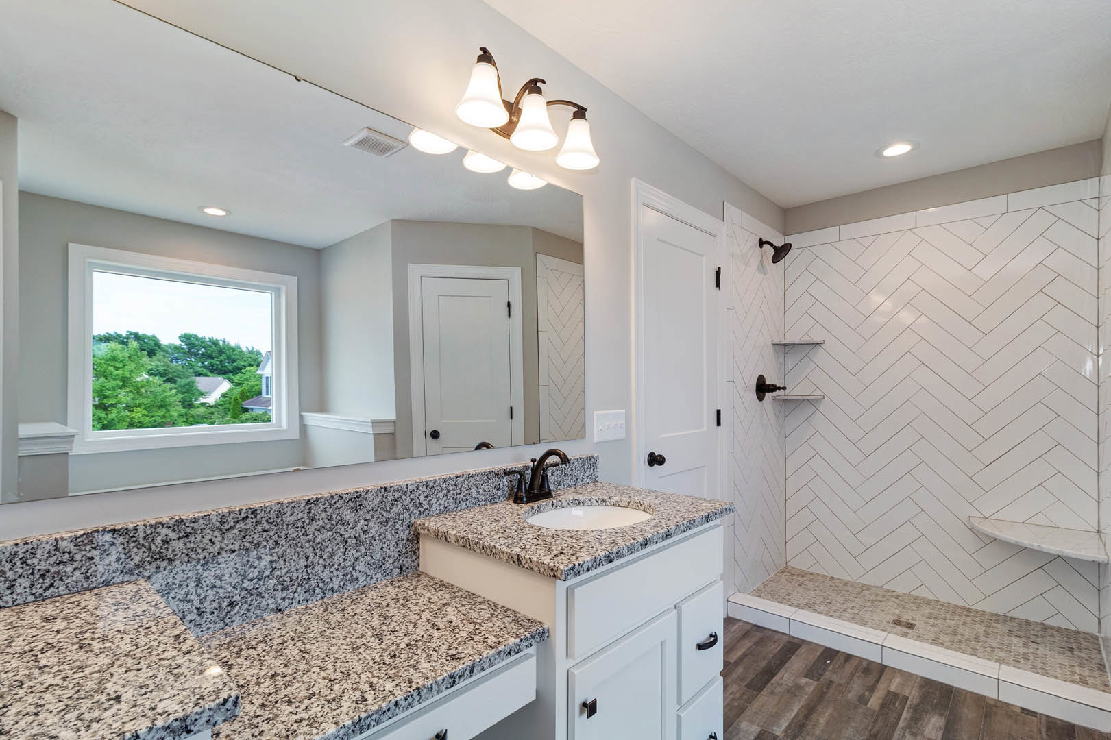 Bathroom featuring a marble countertop with black speckled surface, tile flooring transitioning to wood, glass shower enclosure, white cabinetry with black handles, three-light