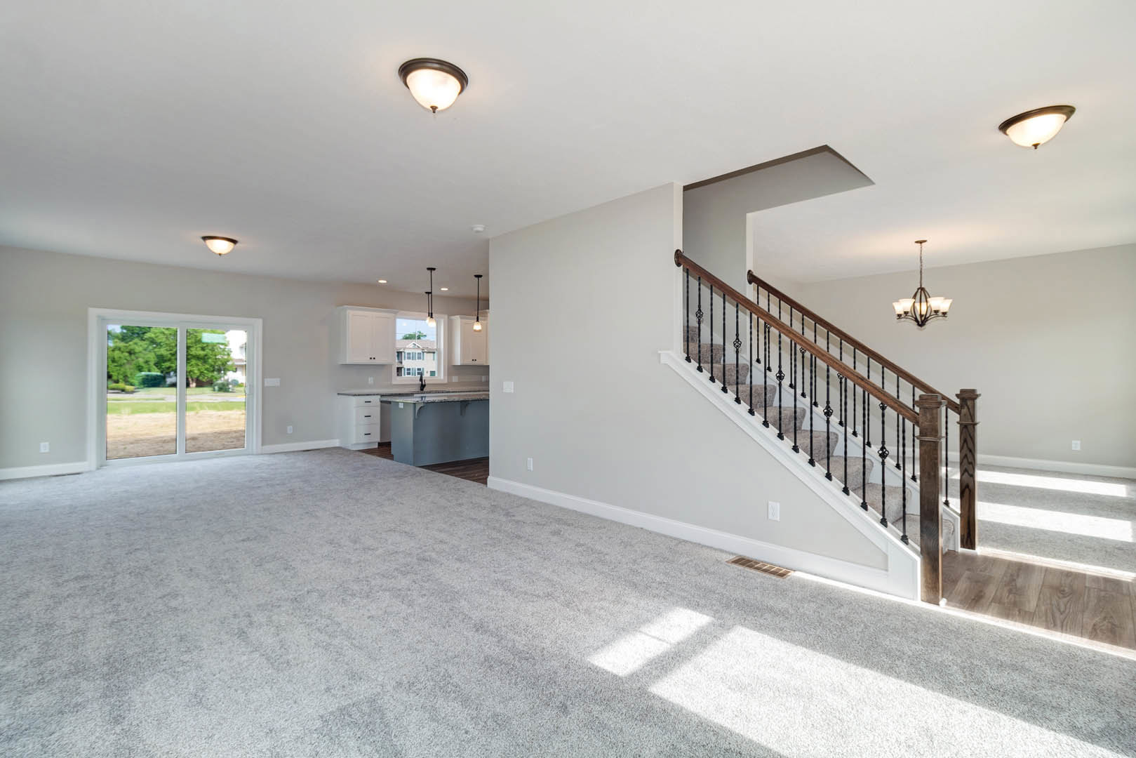 Open-concept interior featuring wood staircase with metal handrail, pendant light fixture, white kitchen cabinetry, large window overlooking green lawn and trees, carpeted room