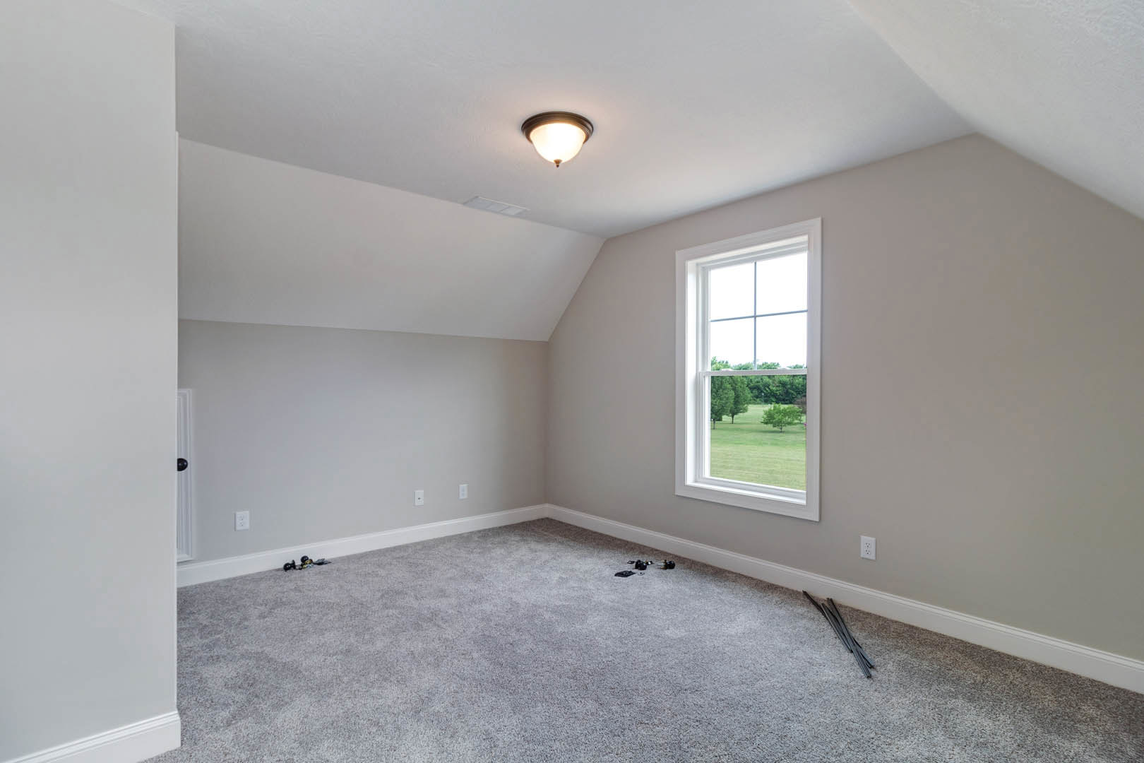 Carpeted room with white walls, large window overlooking green lawn and trees, ceiling light fixture, metal rods and objects scattered on floor