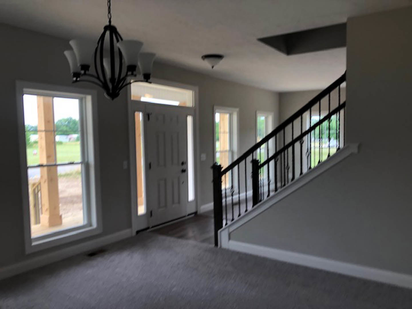 Open staircase with black metal railing, carpeted steps, large window framed by wooden post, modern chandelier overhead, and grey door with black handles in a bright interior