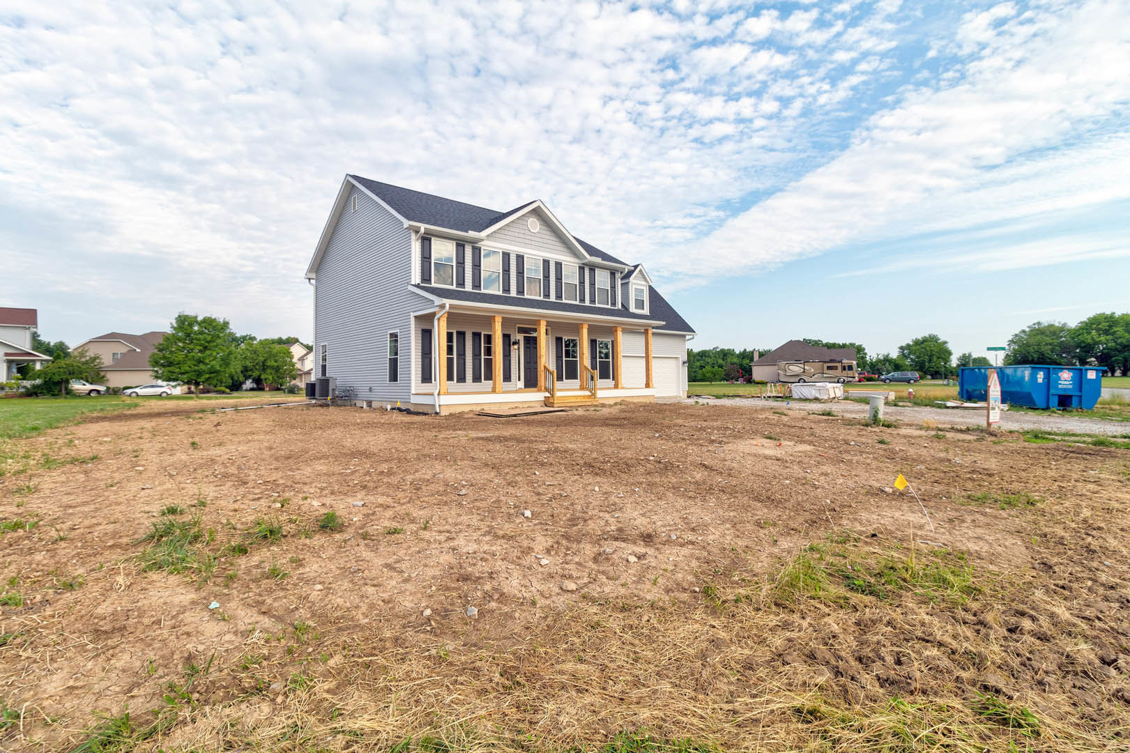 Two-story home with beige siding and white pillars, expansive grassy yard with scattered rocks, blue dumpster near dirt patch, large windows, trees in background, partly cloudy sky