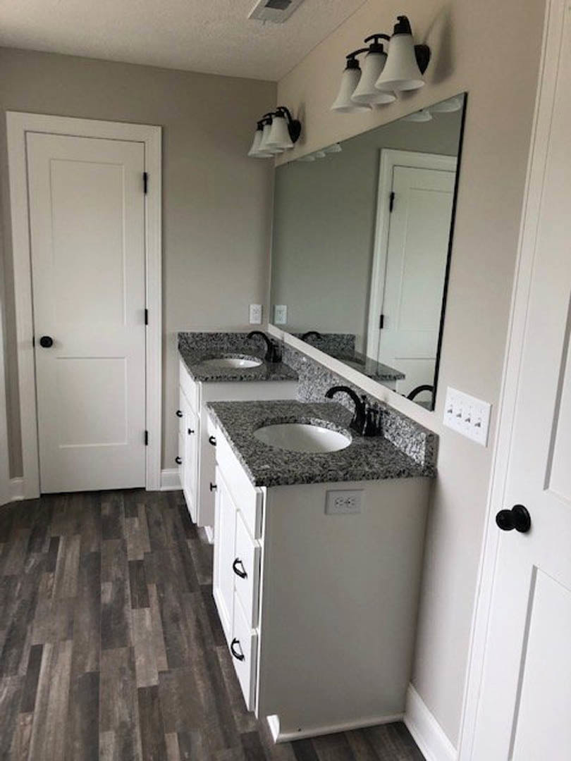 Bathroom with double sinks, wide mirror above, dark wood flooring with white trim, white cabinetry, and overhead group of white lights