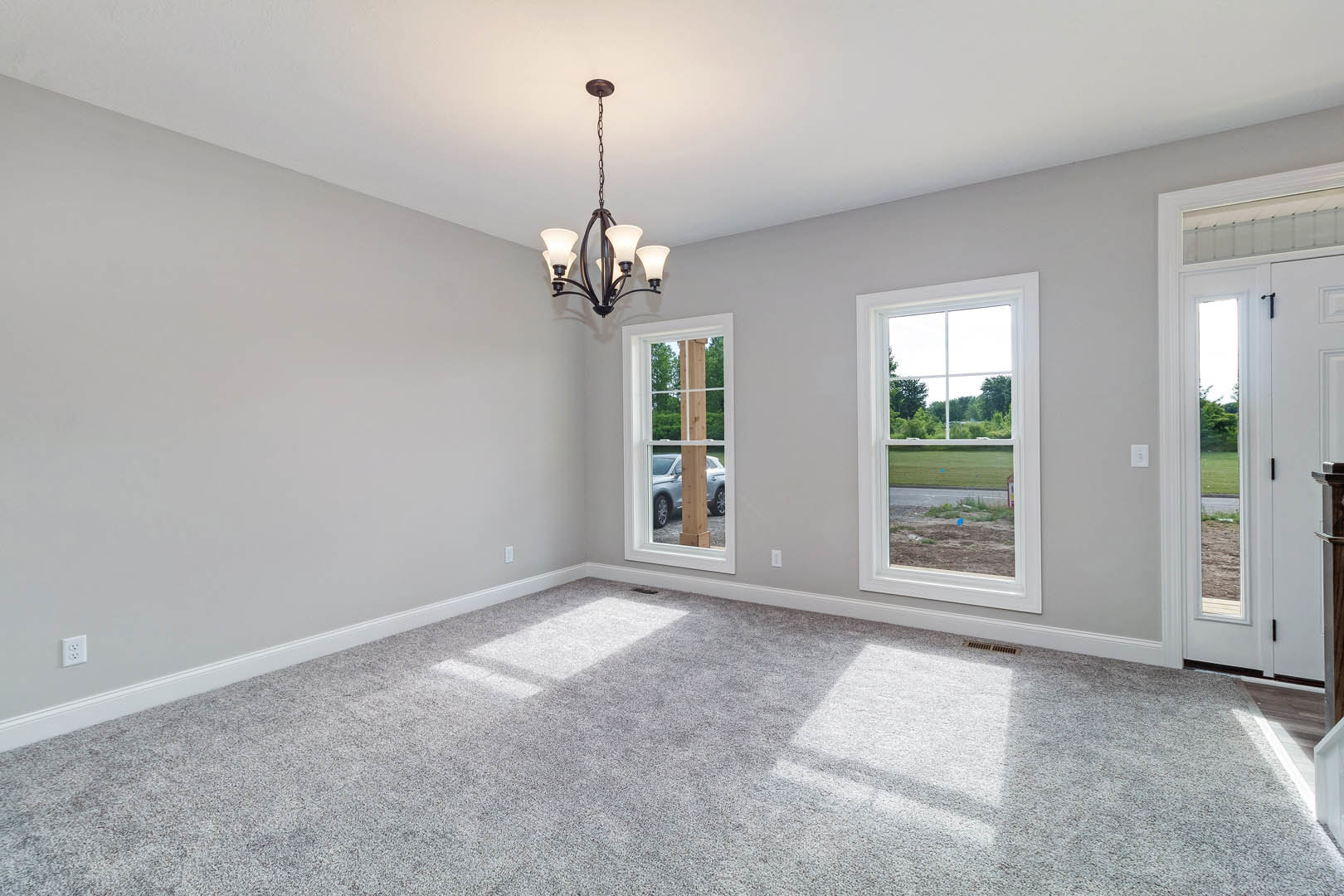 Chandelier hanging from a white ceiling in a carpeted room with large windows, open door leading to green lawn, parked car visible outside