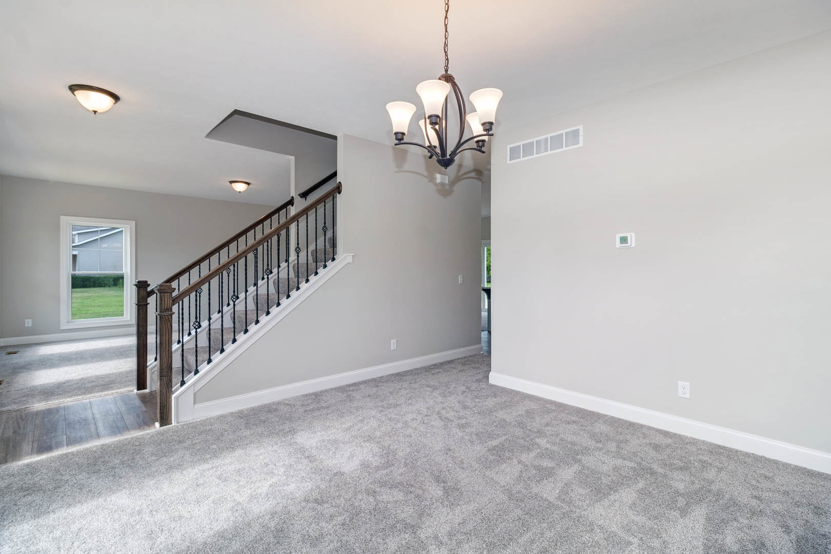 Chandelier with five lights hanging above carpeted floor near staircase with black railing, white rectangular window showing green lawn outside, plaster walls and ceiling