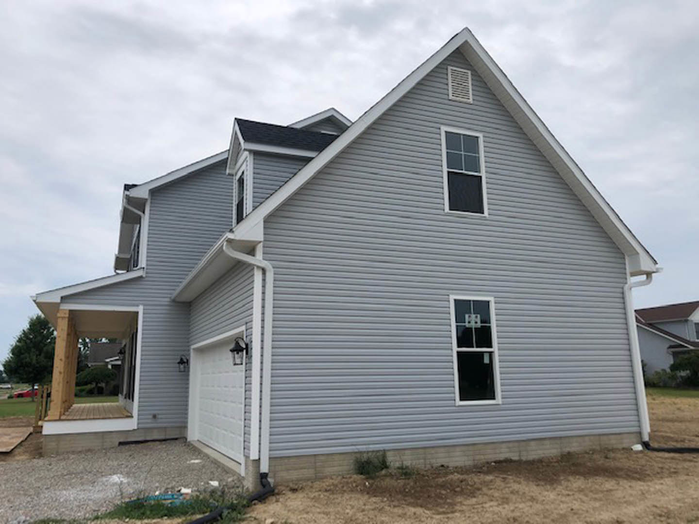 Two-story home with white brick siding, attached garage, white-framed windows, covered porch, and black vent; forest visible in background under partly cloudy sky.