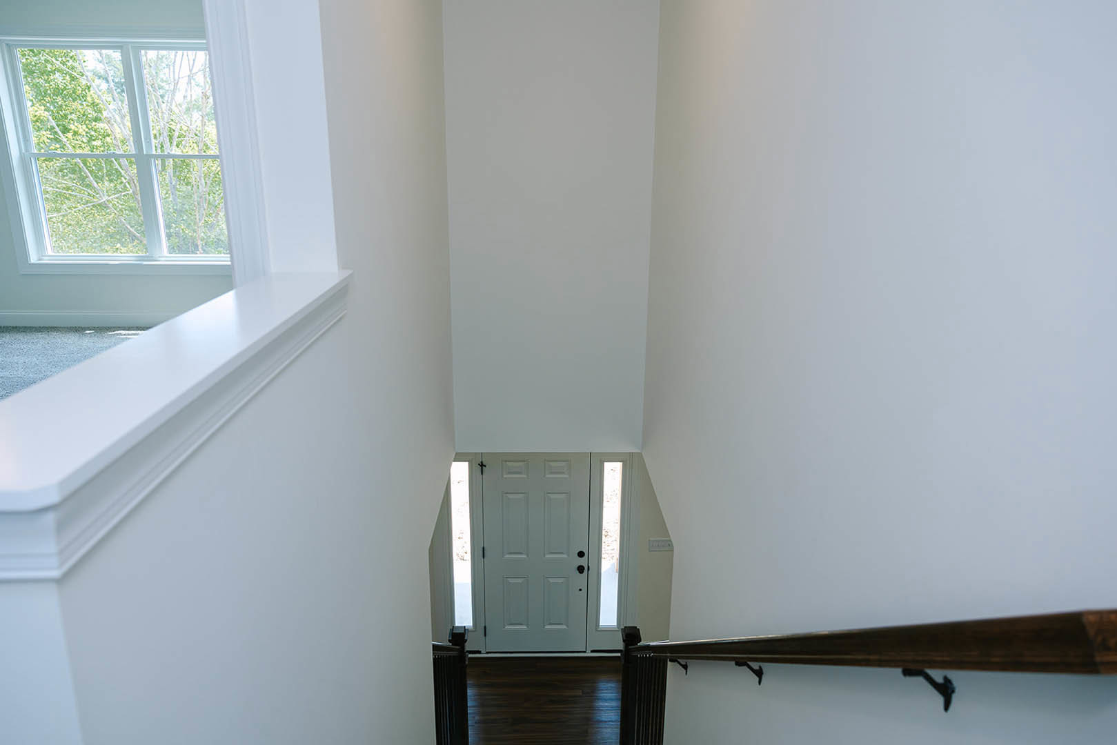 Wood staircase with black metal railing leading to a white door with black handles, adjacent to a large window showing green trees outside, white plaster walls, and light wood