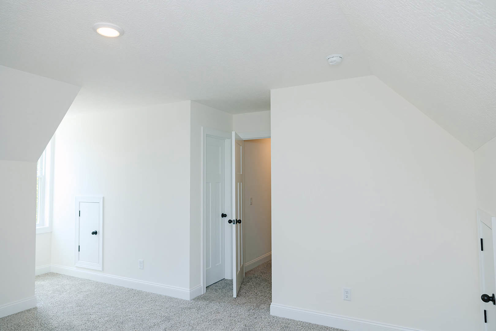 White paneled door with matte black handle partially open against smooth white walls, ceiling light fixture, and light switch visible in modern room with neutral flooring.