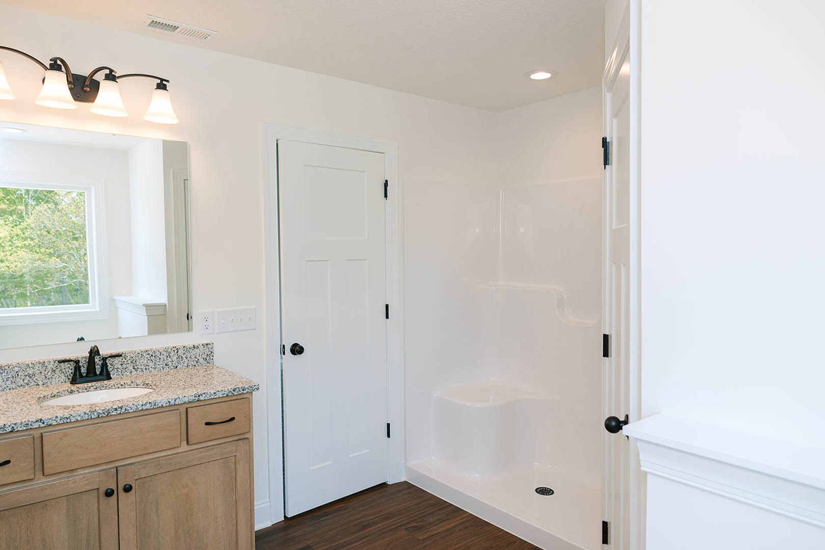 Bathroom with white tile shower, glass door, white vanity with undermount sink, black faucet, marble countertop, two-light wall fixture, white door with black knob, window showing
