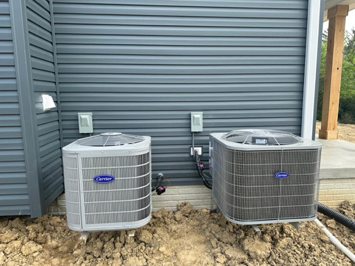 Two white air conditioning units on concrete pad beside exterior wall, wooden post with drilled hole, waste container partially visible, light switches mounted on wall