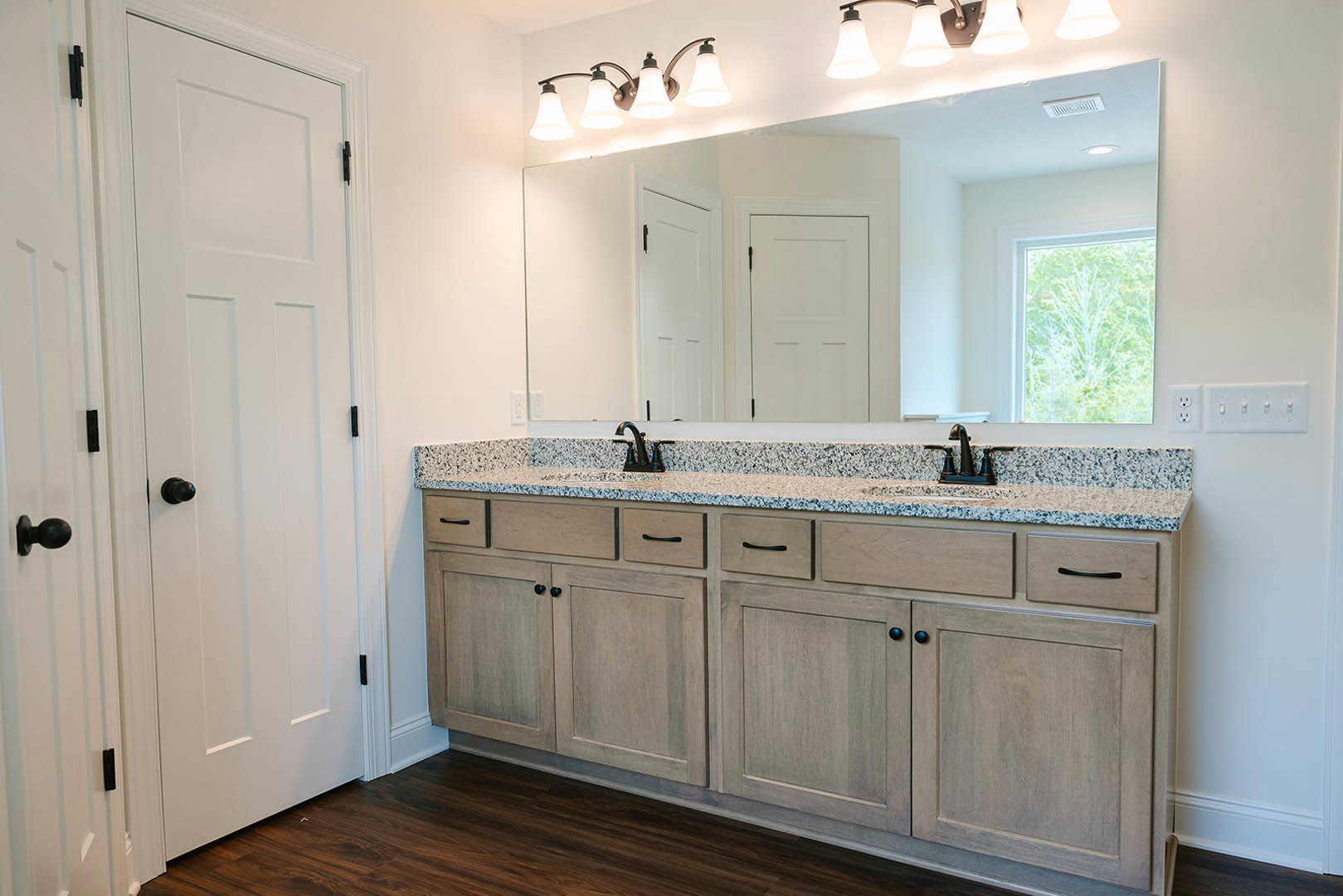 Bathroom with marble double vanity, large framed mirror, white cabinetry with black hardware, white door, window overlooking trees, and tiled walls.