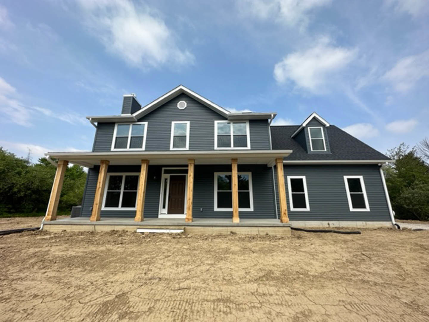 Two-story house with white-framed windows, covered porch, and dirt yard in front, surrounded by trees under a cloudy sky