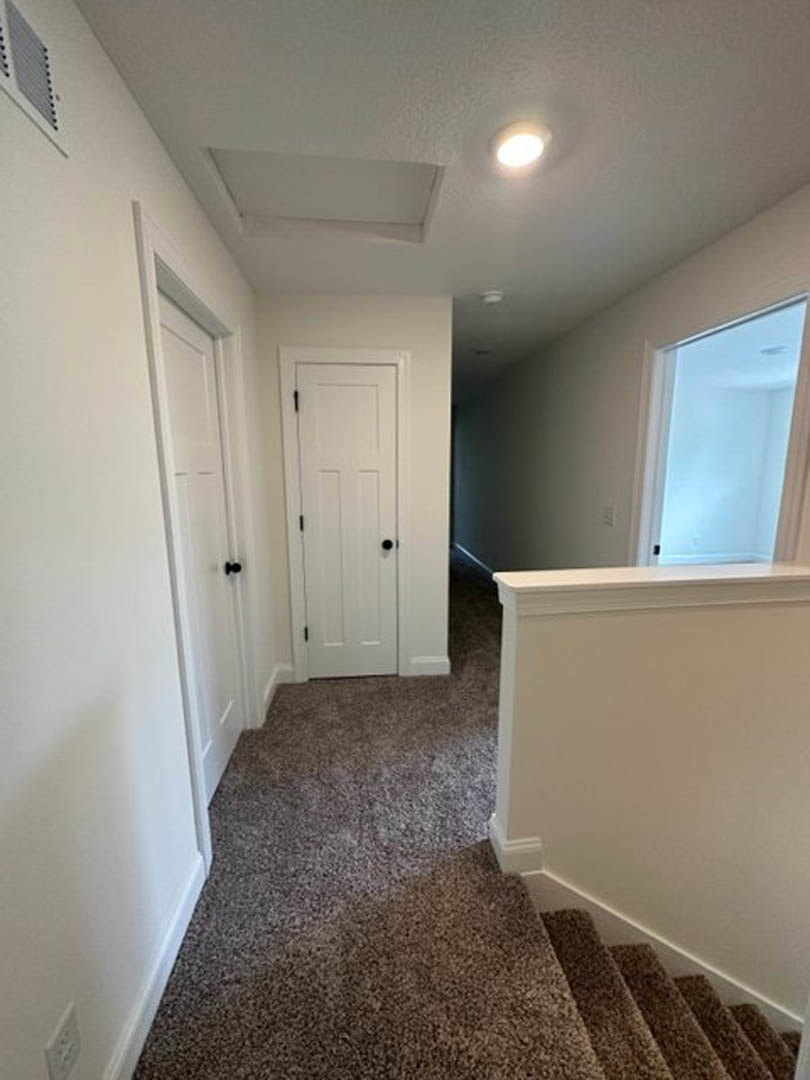 Carpeted hallway with white walls, multiple white doors featuring black knobs, ceiling light fixture, and a carpeted staircase visible along one side