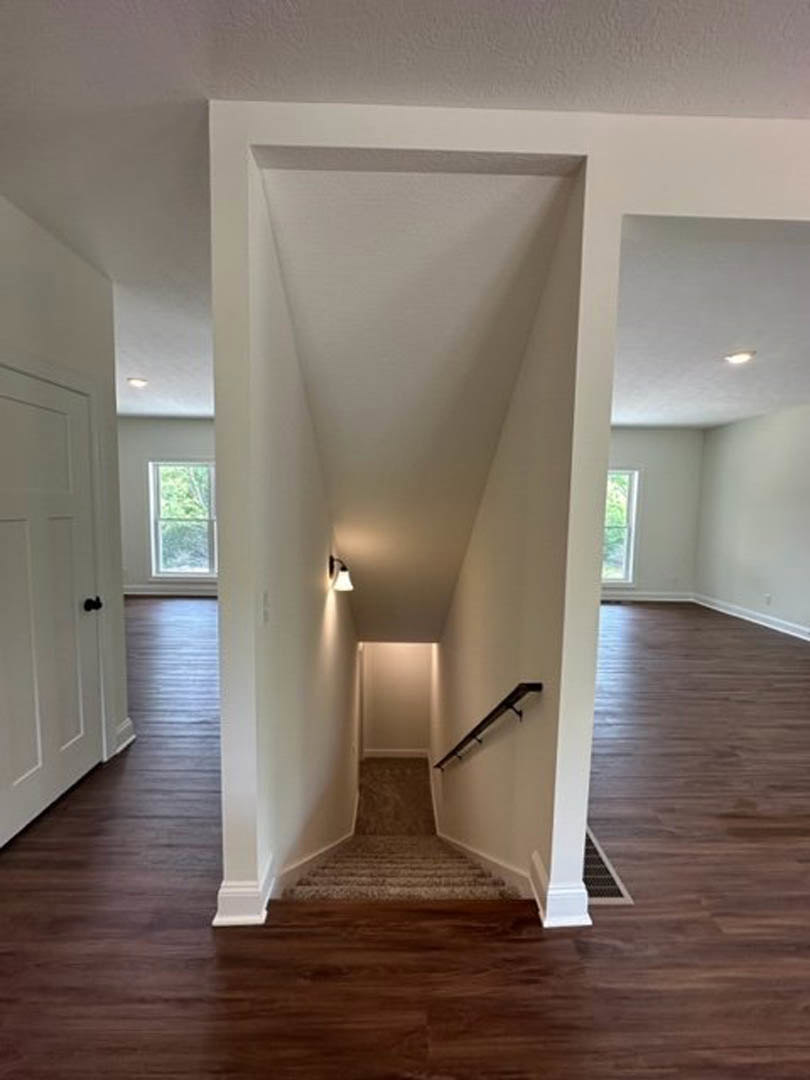 Hardwood staircase with white risers and wooden handrail, hallway with laminate flooring, recessed ceiling lights, white plaster walls, and white door featuring black knob