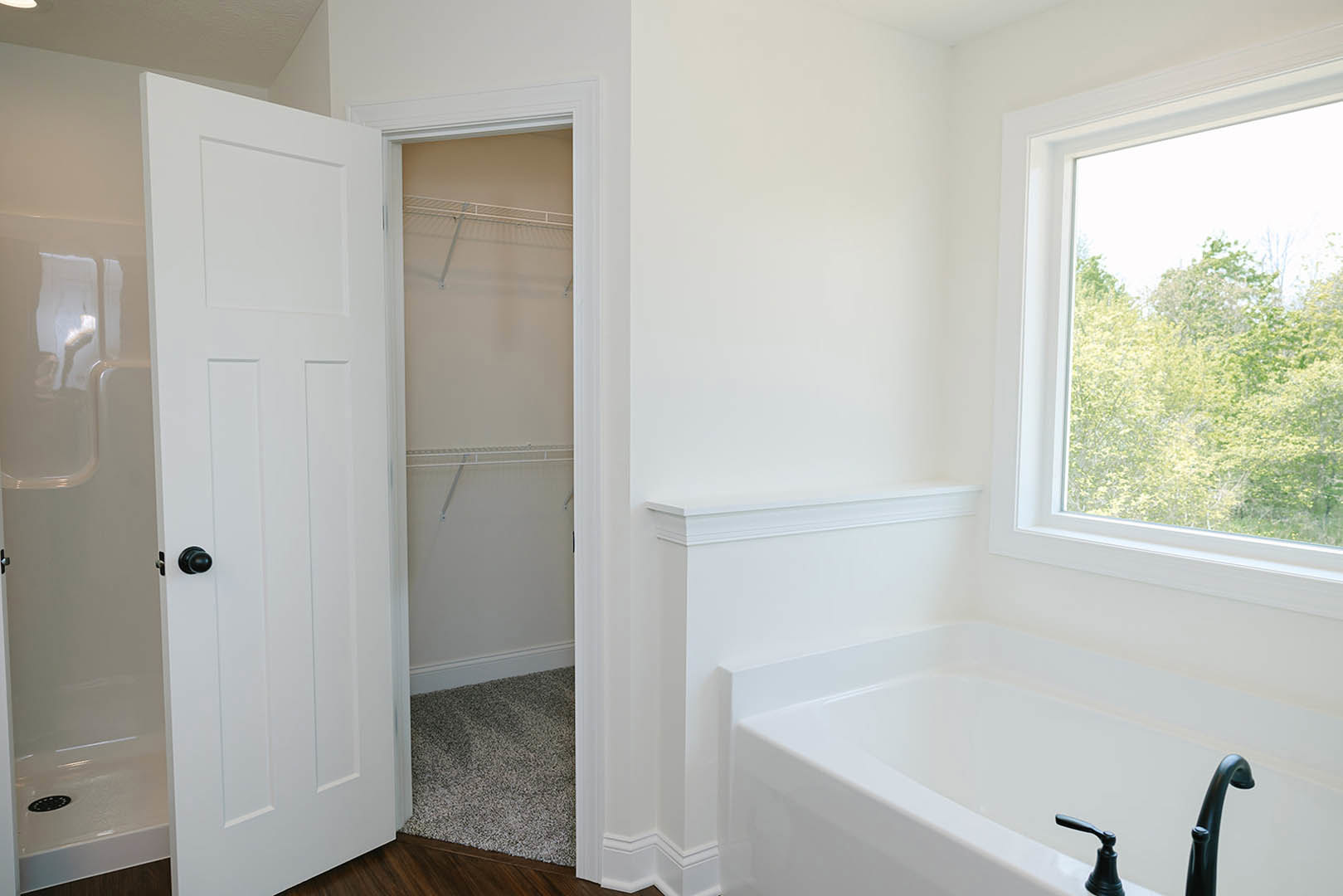 White bathroom with freestanding bathtub, open white door with black knob, white walls and trim, window overlooking trees, light carpet flooring