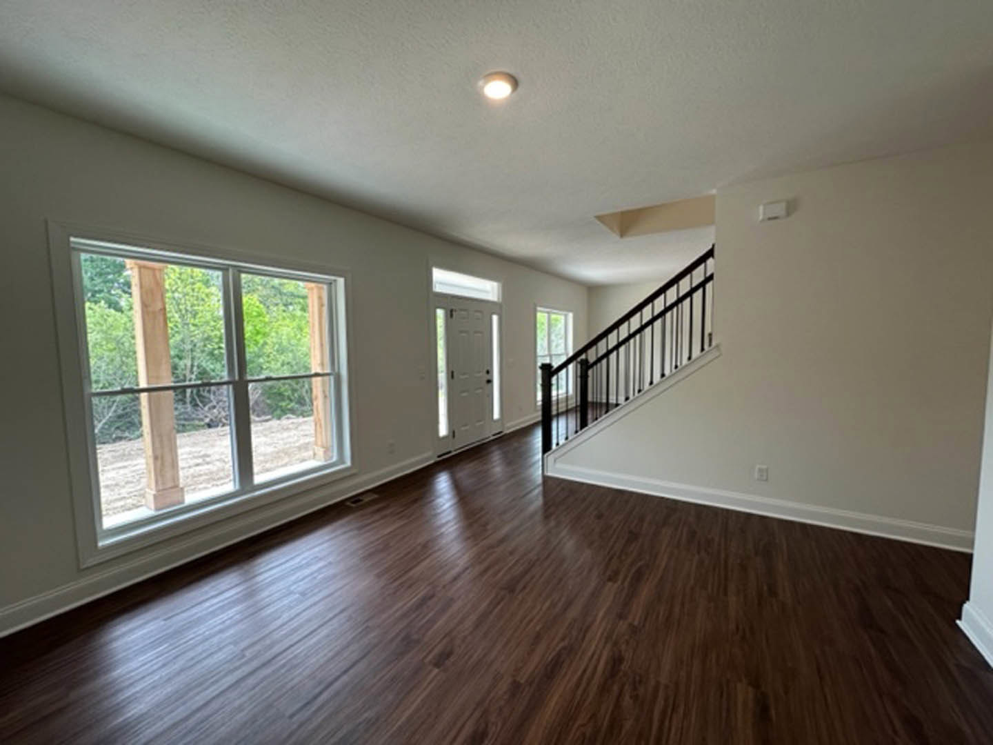 Hardwood floored hallway featuring a staircase with black railing, ceiling light fixture, white door with window, and large window overlooking trees.