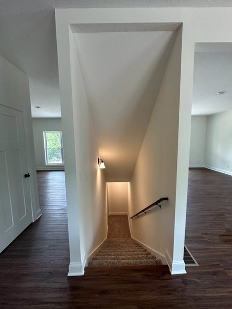 Wood staircase with wall-mounted lights, white door with black knob at the landing, metal rod with hooks, laminate flooring, window overlooking greenery.
