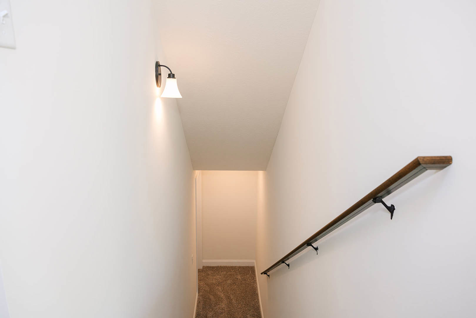 Carpeted staircase with wooden handrail, white plaster walls, wall-mounted light fixture, and hooks along the rail.