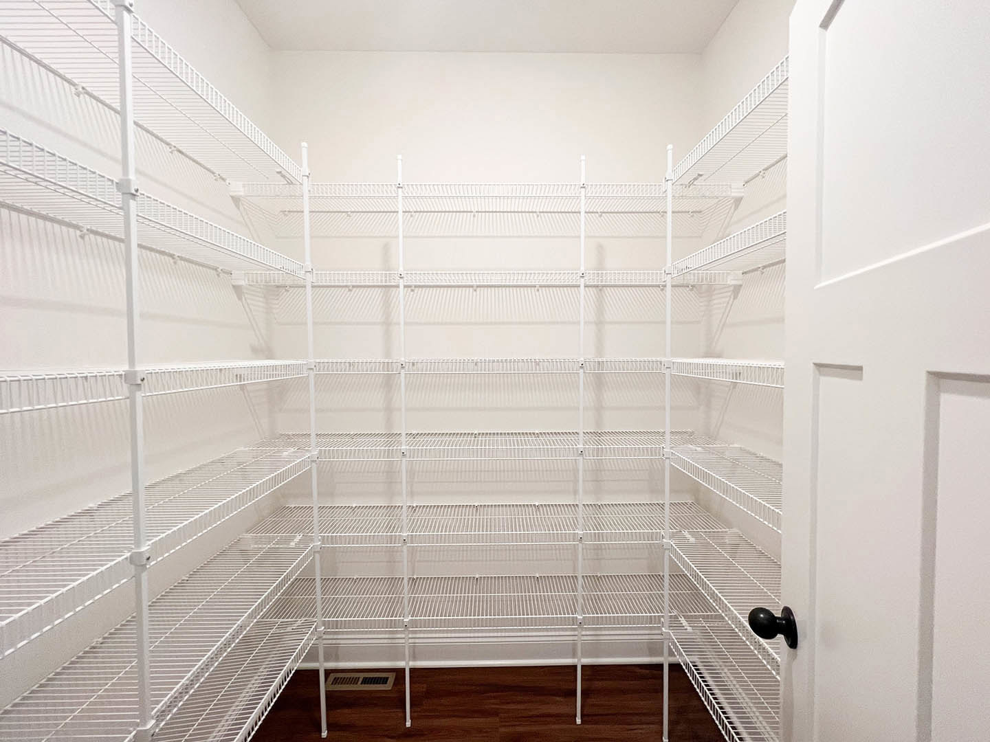 Large white built-in shelves in a spacious room with light wood flooring, white closet door featuring a black knob, white metal pole lamp, and a decorative white wire cage placed