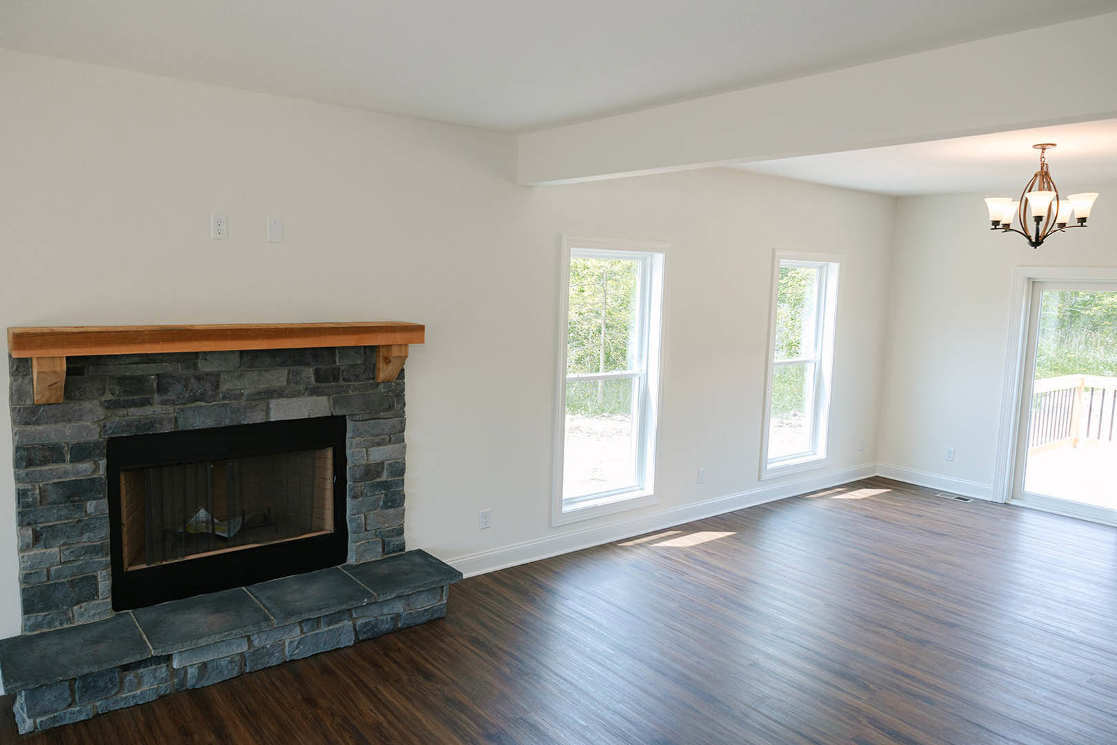 Living room with stone fireplace featuring metal grill and wood mantel, hardwood flooring, chandelier, large window overlooking trees.