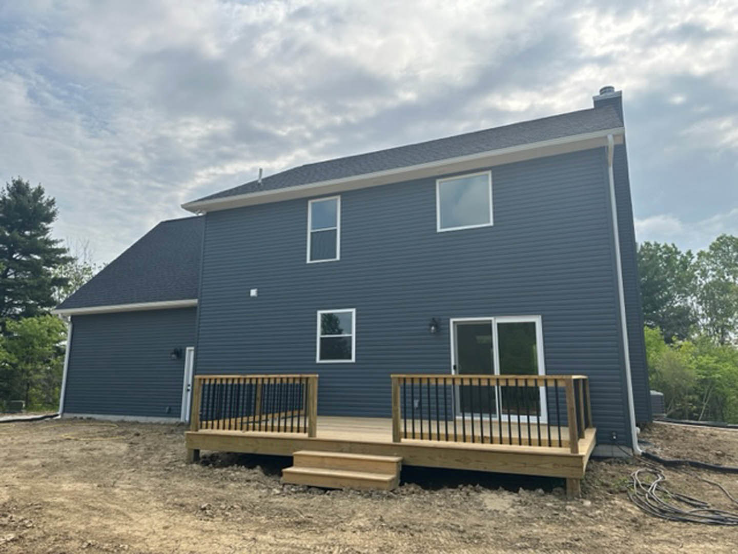 Two-story house with gray siding, wooden deck featuring white railing, fenced backyard, large windows with white frames, and patio area surrounded by trees under a cloudy sky
