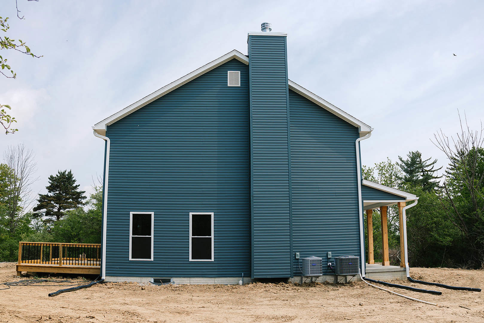 Blue siding house with white trim, covered front porch featuring wooden deck and black metal railings, brick chimney, large window with black frame, wall vent, and outdoor air