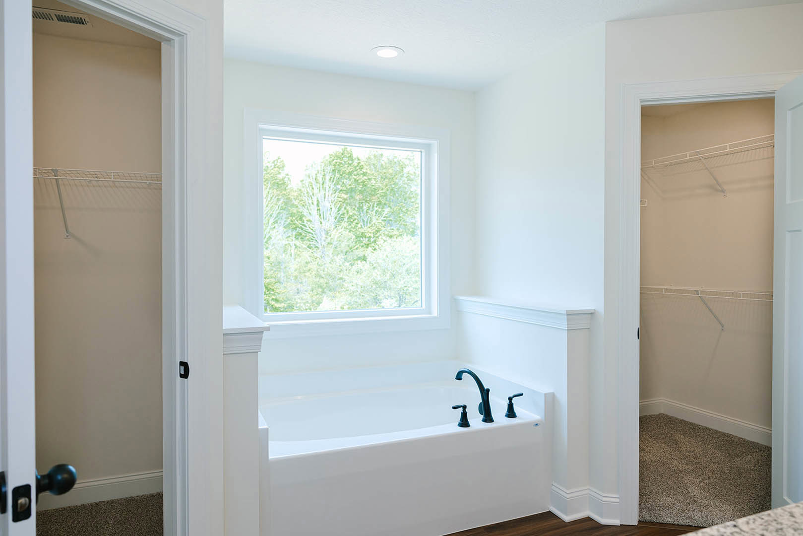 Freestanding white bathtub with matte black faucets beneath a large window showing green trees, white walls, and curtain rod in modern bathroom