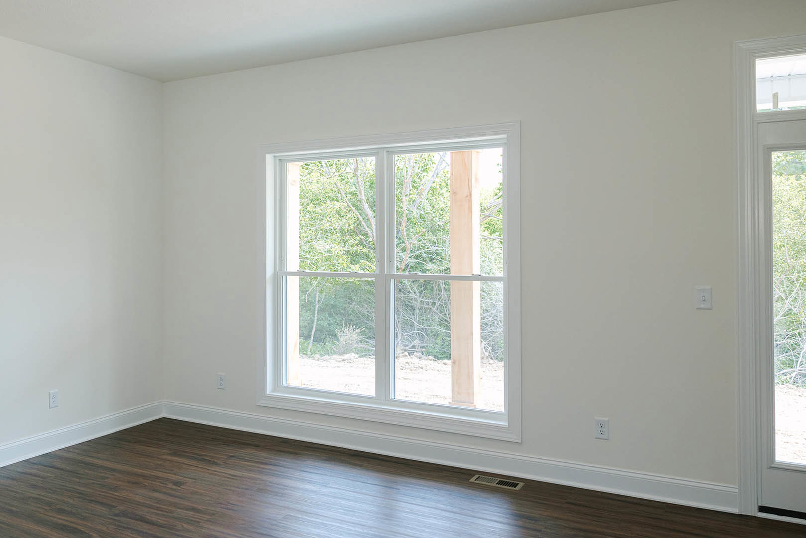 Sunlit room featuring a large window with white frame, hardwood flooring, white walls, and views of green trees outside.