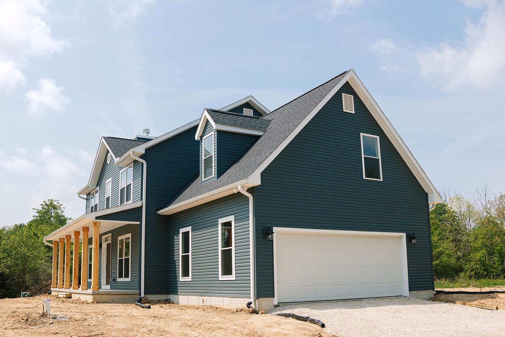 Two-story home with blue roof, white-framed windows, attached garage with paneled door, wooden pillars along covered entry, surrounded by tall trees and cloudy sky