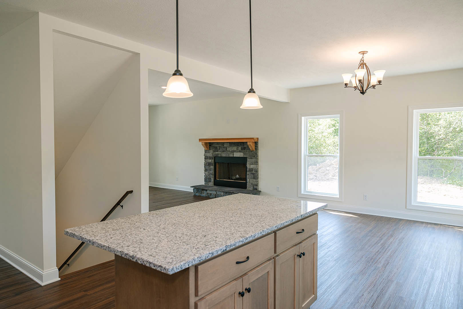 Kitchen island with wood cabinetry and stone countertop, fireplace with wood mantel, large window showing trees, five-light ceiling fixture, hardwood flooring