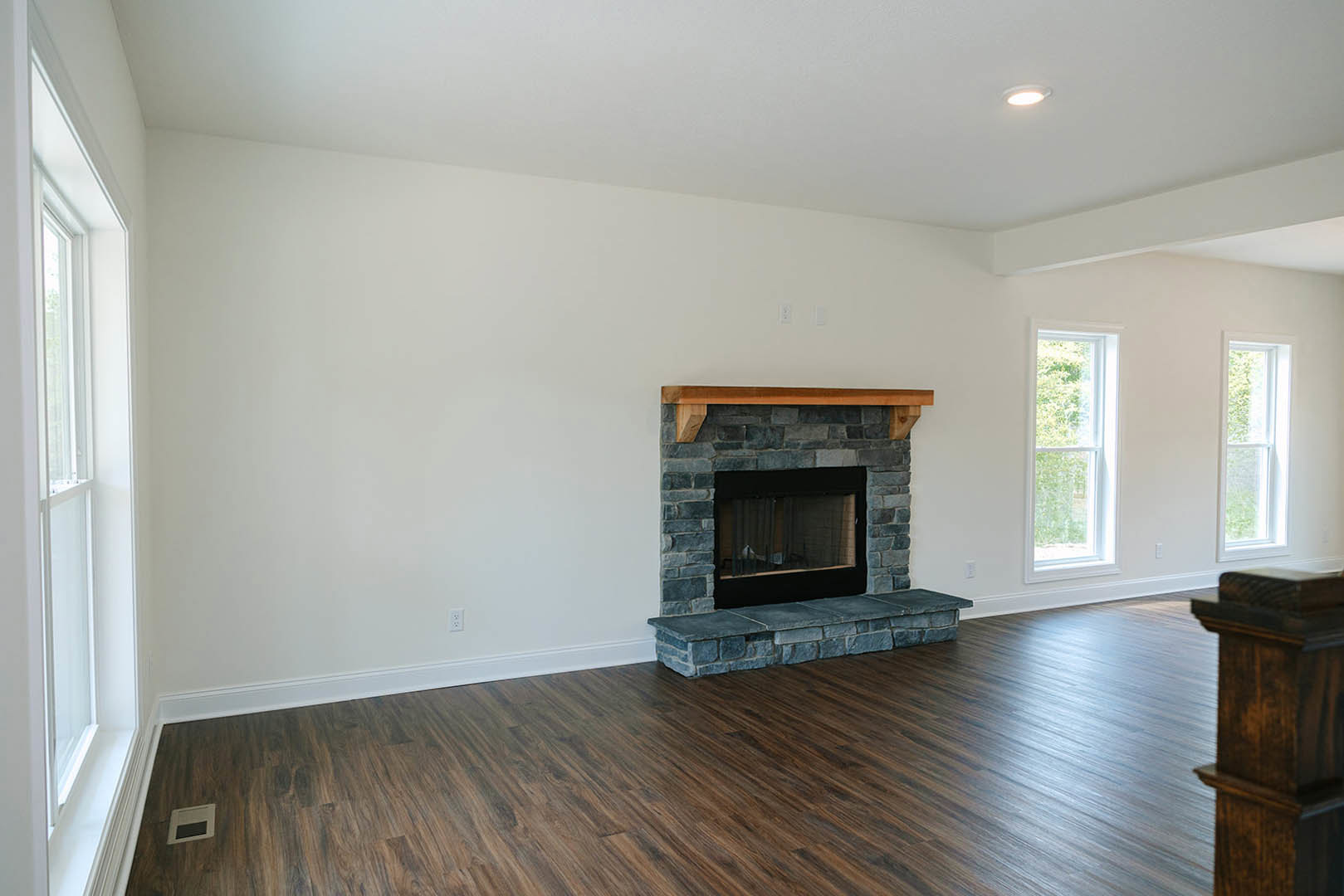 Stone fireplace with wood mantel set against a textured stone wall, black-framed window to the side, hardwood flooring throughout the room.