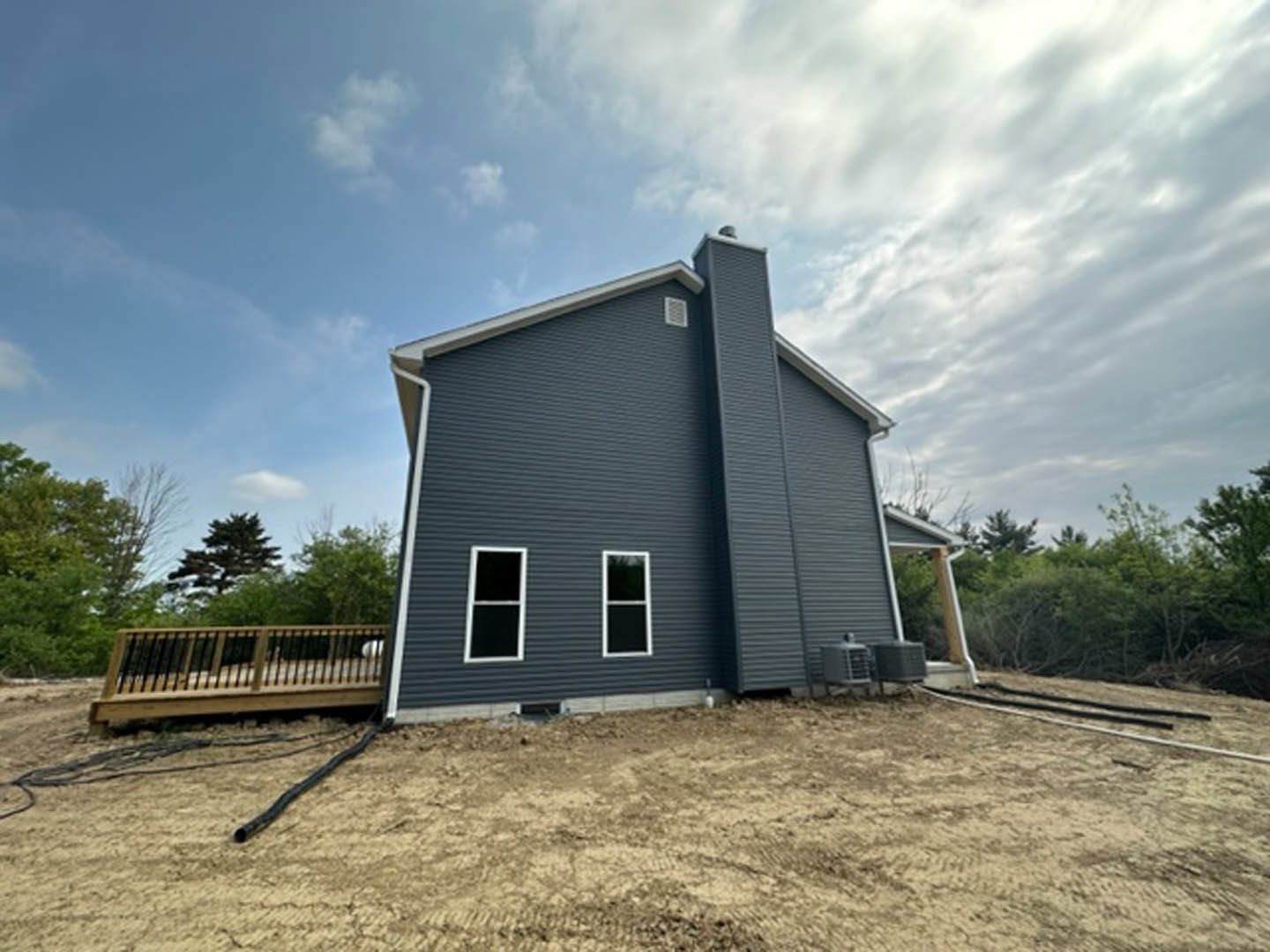 Two-story house with light siding, white-trimmed windows, attached wooden deck, fenced backyard, and chimney, surrounded by trees and patchy dirt ground.
