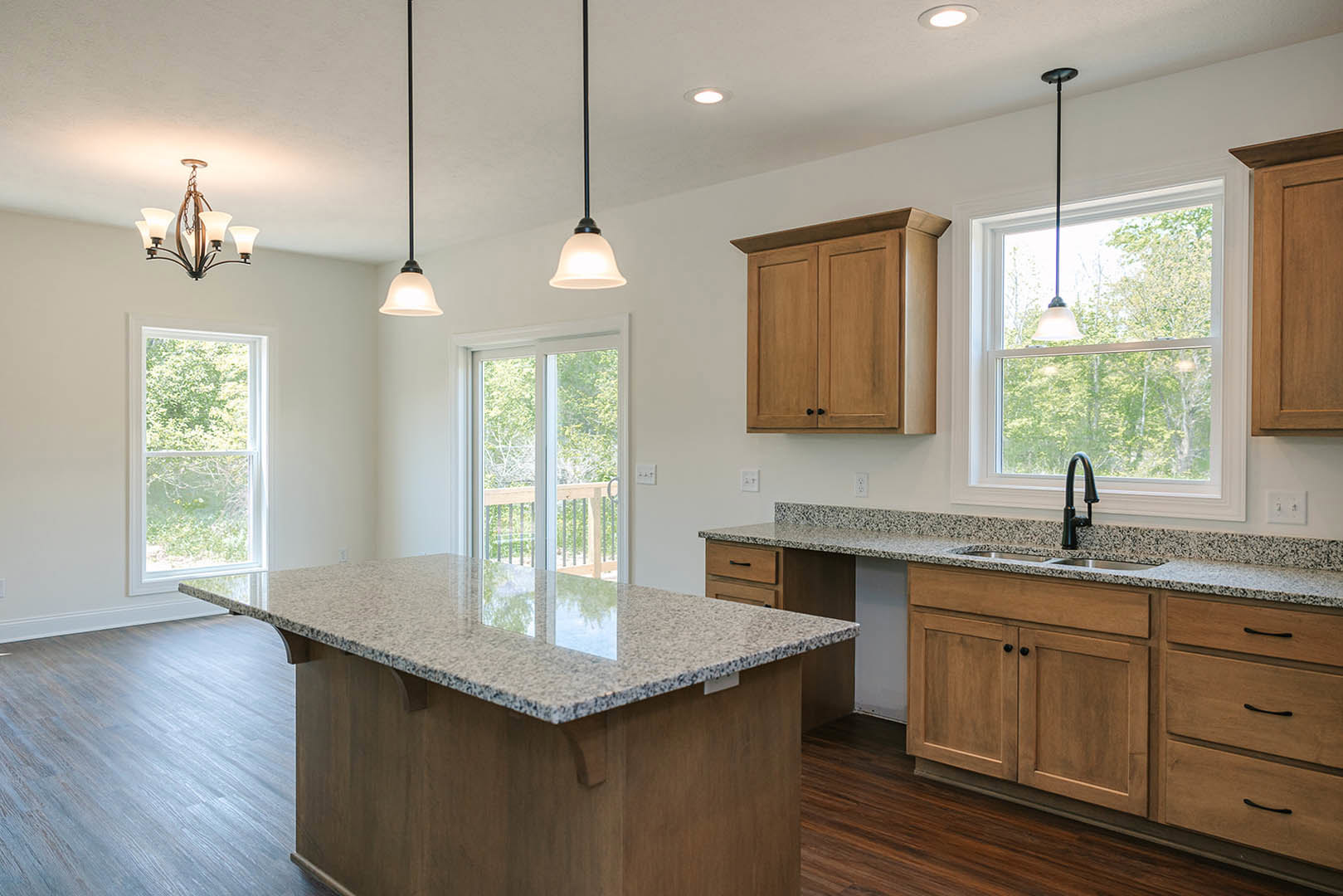 Granite countertops and wood cabinets with black handles, stainless steel sink and tap, white walls with electrical outlets, window overlooking trees, chandelier overhead