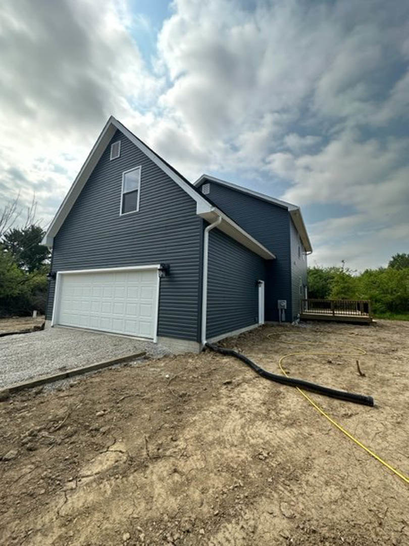 Two-story house with light siding, attached garage with white door, garden hose coiled on driveway, wooden fence and trees in background, single window on front facade, black hose