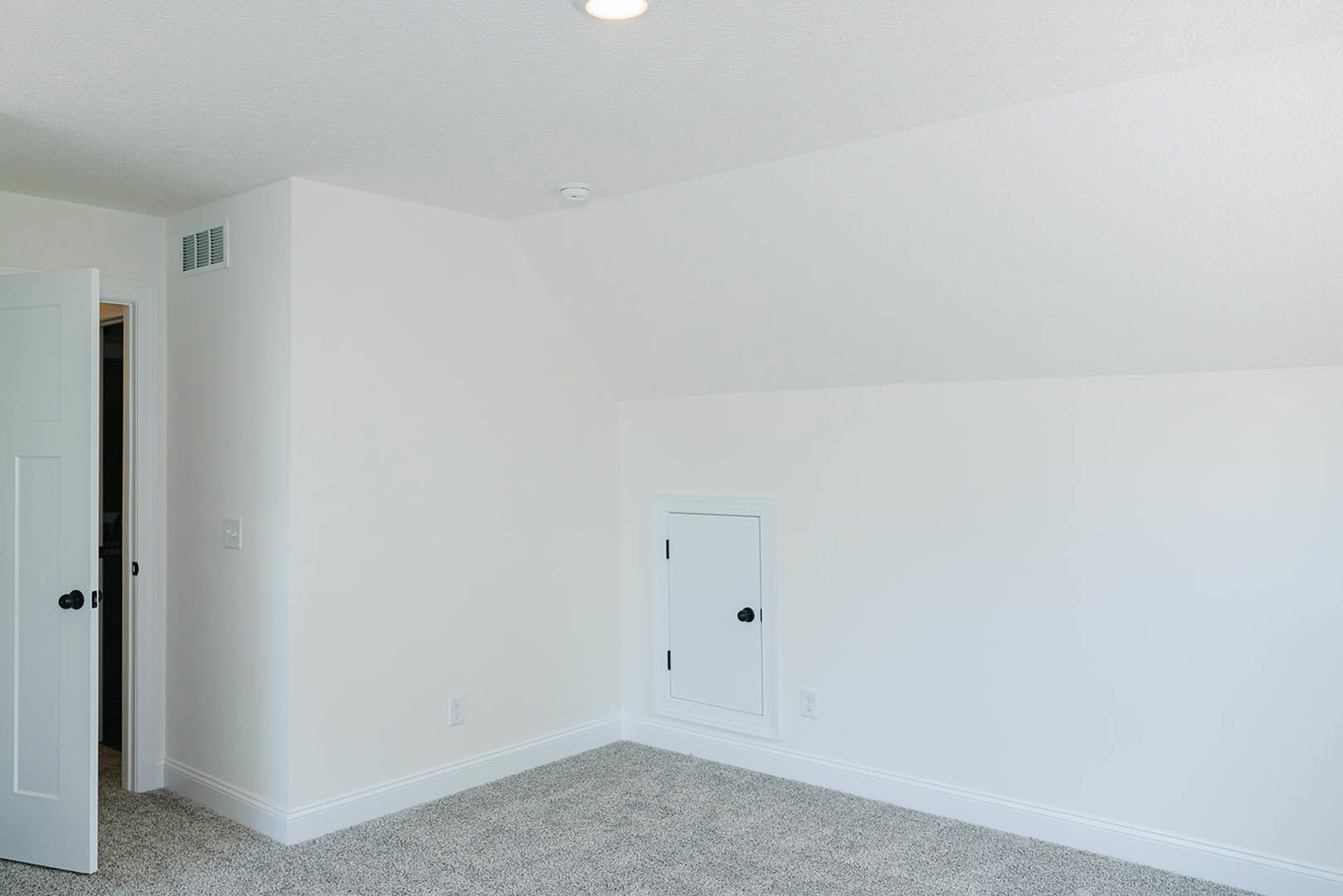 White-painted room with white door featuring black knob, carpeted floor, wall vent, and smooth plaster walls.