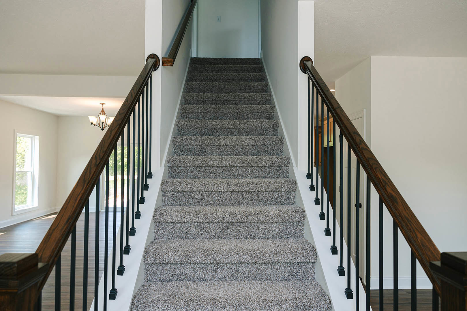Grey carpeted staircase with black metal railings, white framed window on the landing, and a modern chandelier overhead.