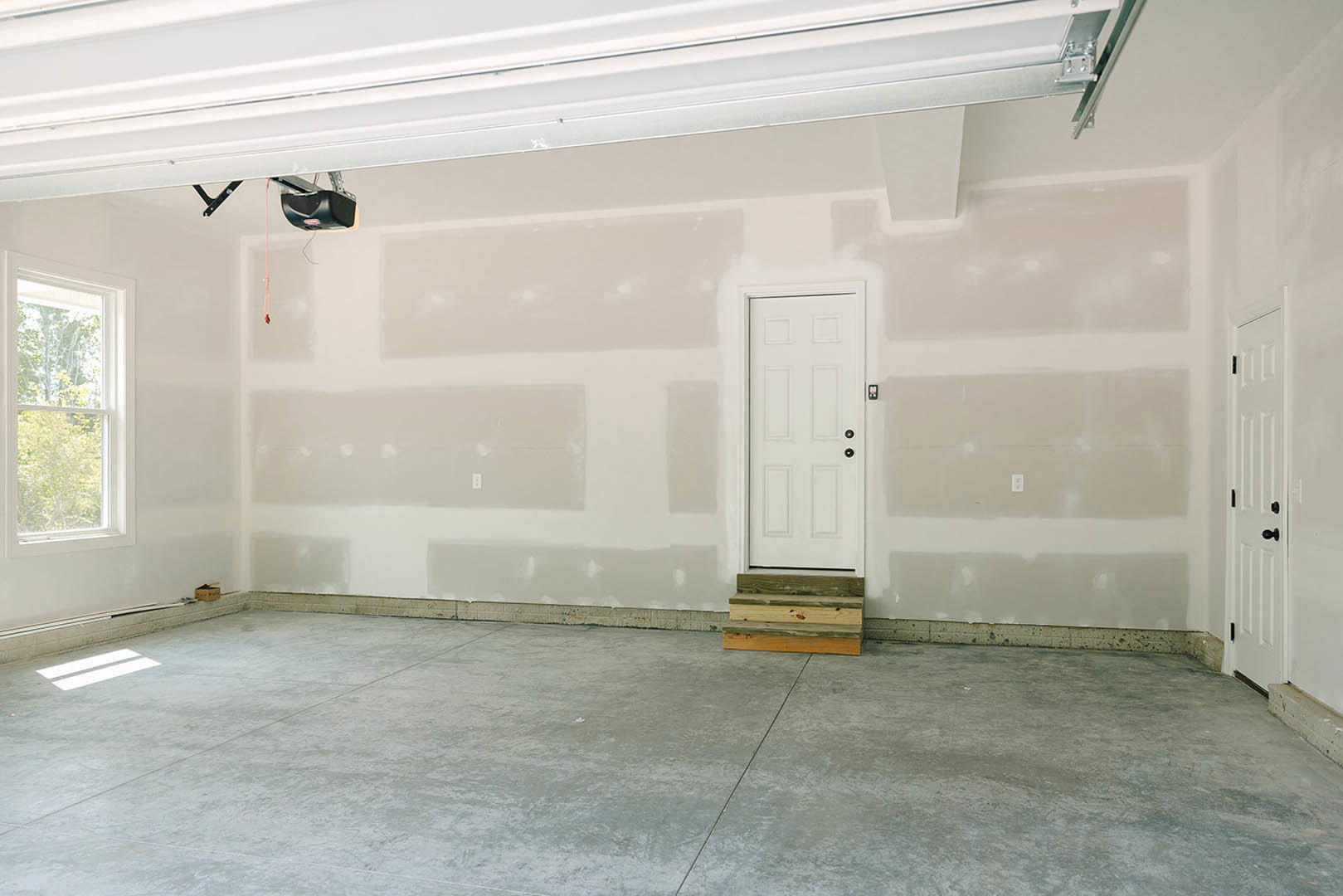 White door with black knobs, concrete floor featuring a wooden step, plaster walls, window overlooking trees, close-up of wood plank and white ceiling.