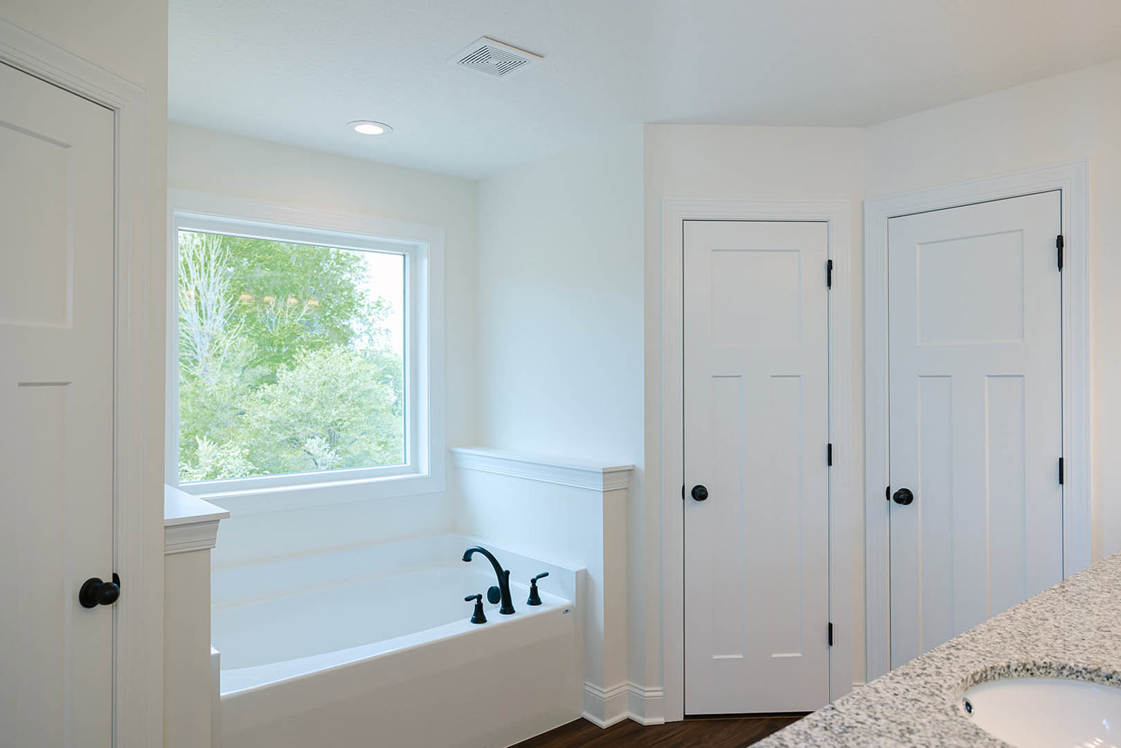Bathroom with freestanding white bathtub under a window showing trees outside, white door with black knobs, ceiling vent, close-up of white sink, black faucet fixtures