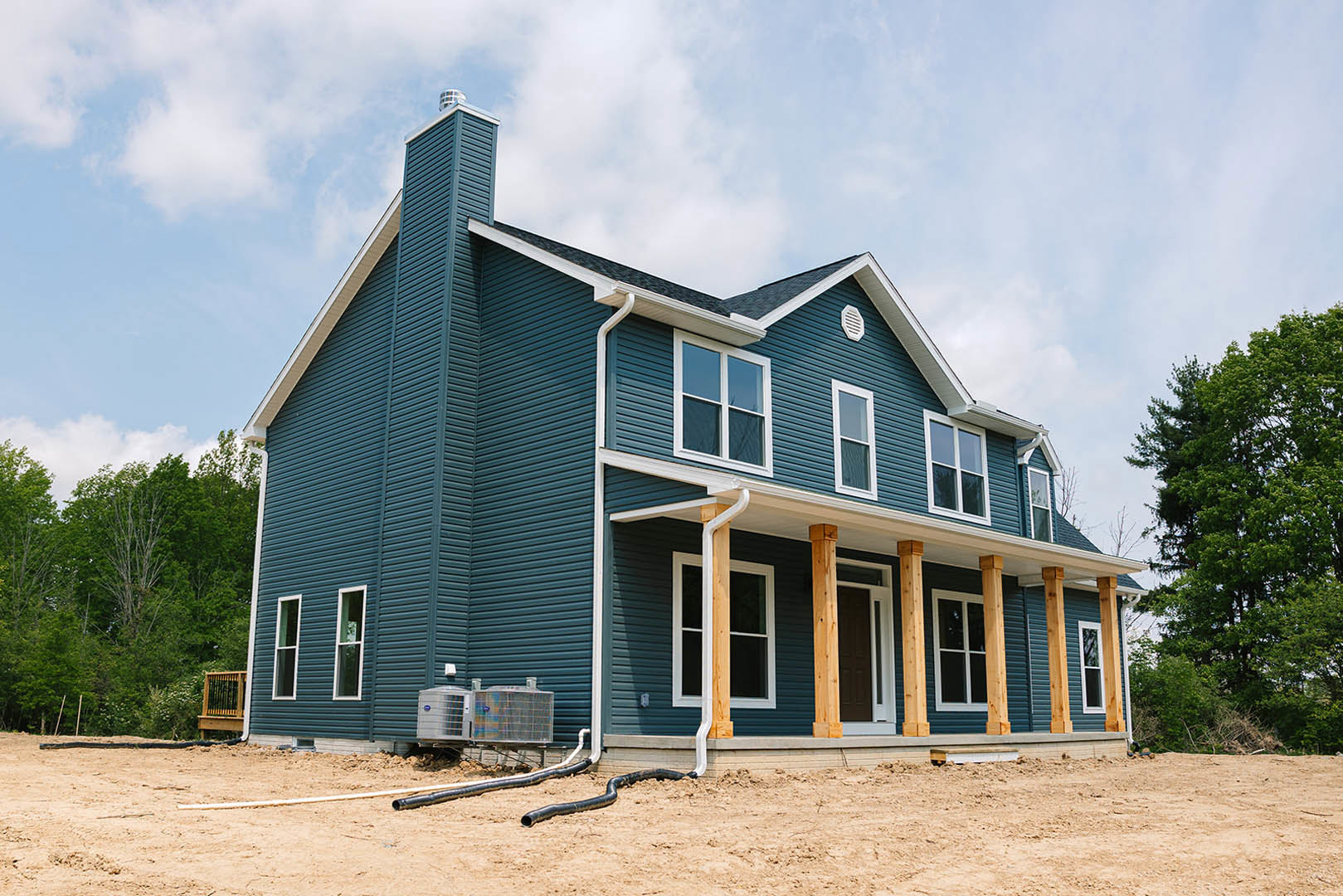 Partially built house exterior with white-framed windows, wooden deck and railing, leafy trees surrounding property, under clear blue sky