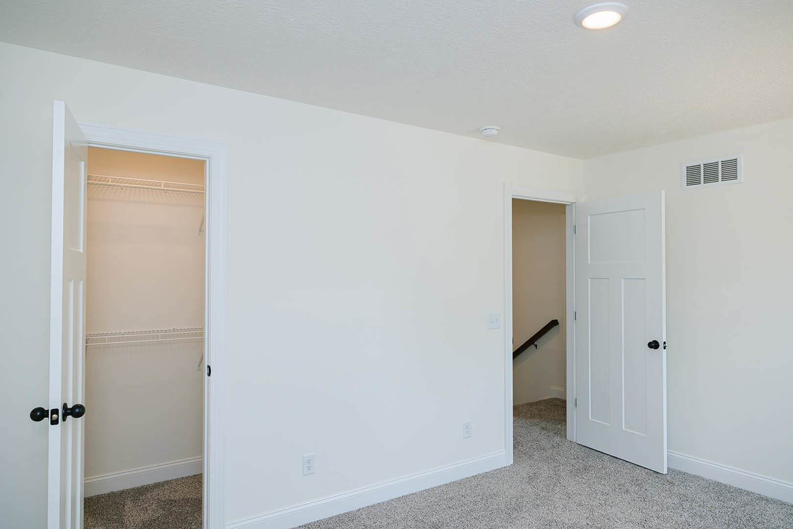 White-walled room with open closet door featuring black knobs, ceiling light fixture, and visible floor vent