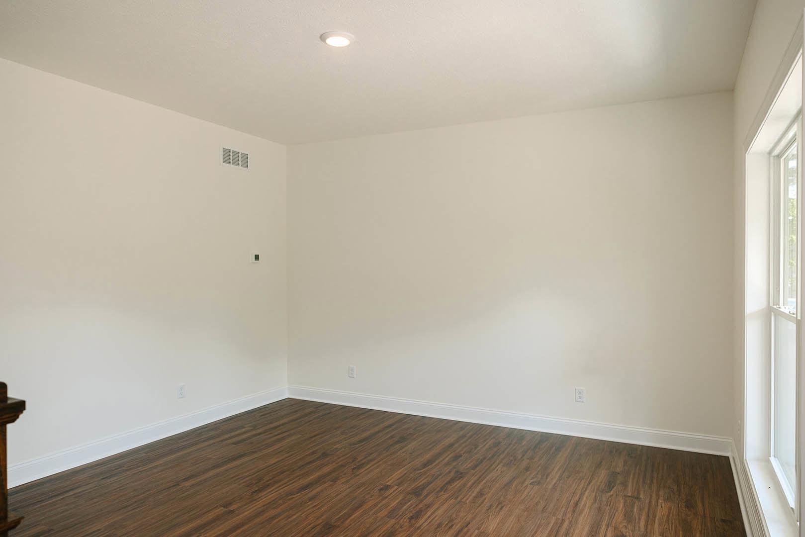 Wood floor with white baseboards, white plaster walls, brown-framed doorway, ceiling light fixture, and row of wall vents in a residential interior.