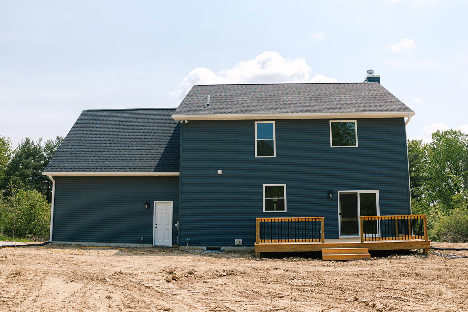 Wooden deck with railings attached to a partially constructed house, white door with black knobs, white-framed window reflecting blue sky, exterior surrounded by trees and clouds.