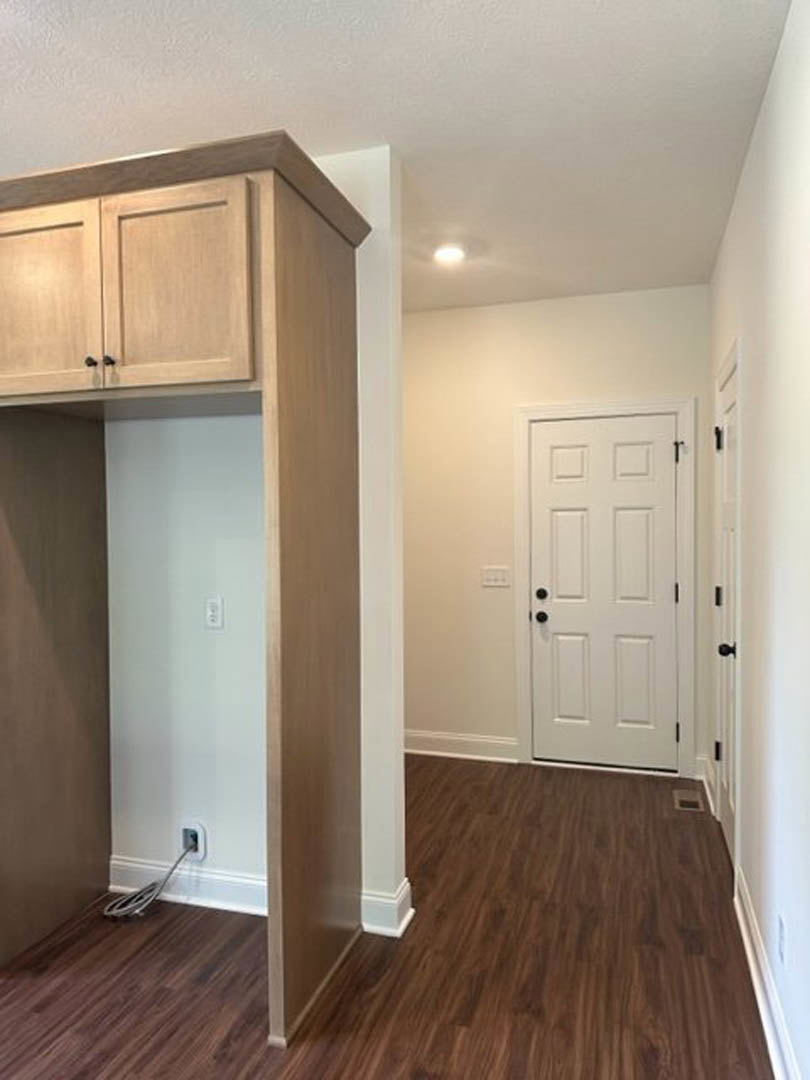 White door with black knobs, wood floor with floor vent, white wall featuring light switch and electrical outlet with cord plugged in