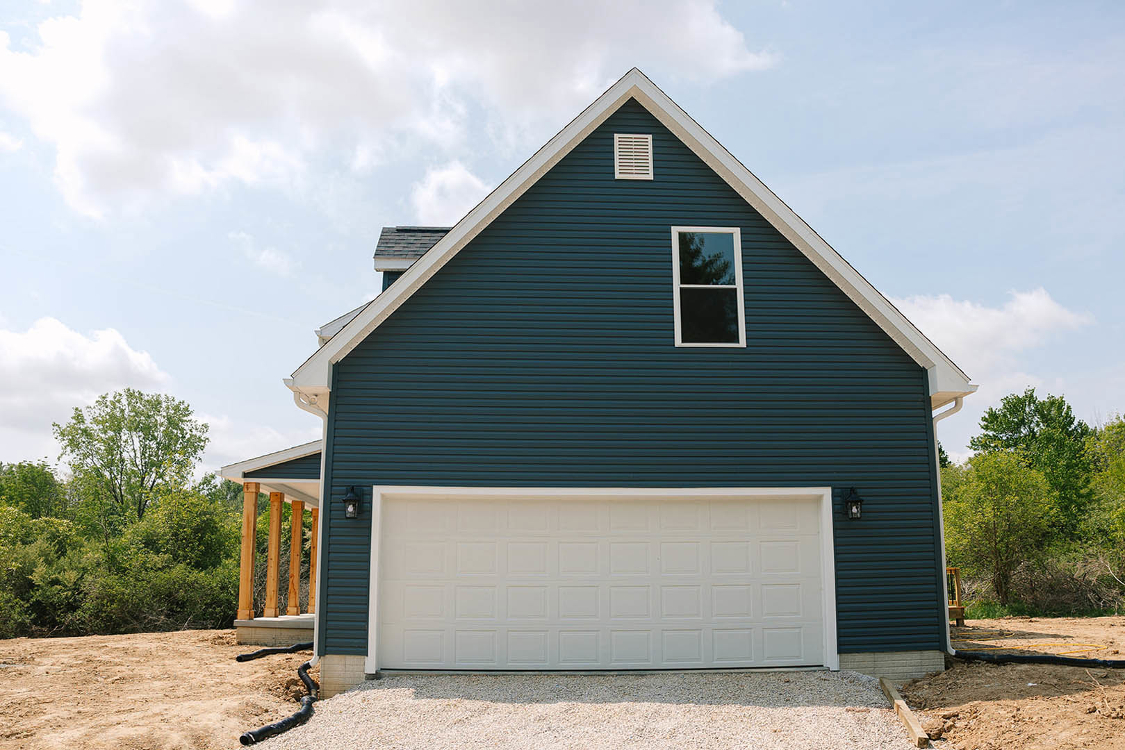Blue siding custom home with attached garage, white vent, large window reflecting leafy tree, gravel driveway, and green landscaping.