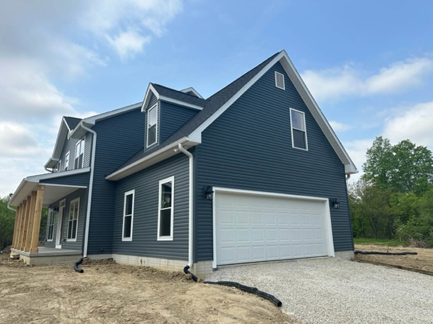 Two-story home with white siding, wooden pillars supporting a white roof over the entry, large garage door, paved driveway, white-framed windows, and trees in the background under