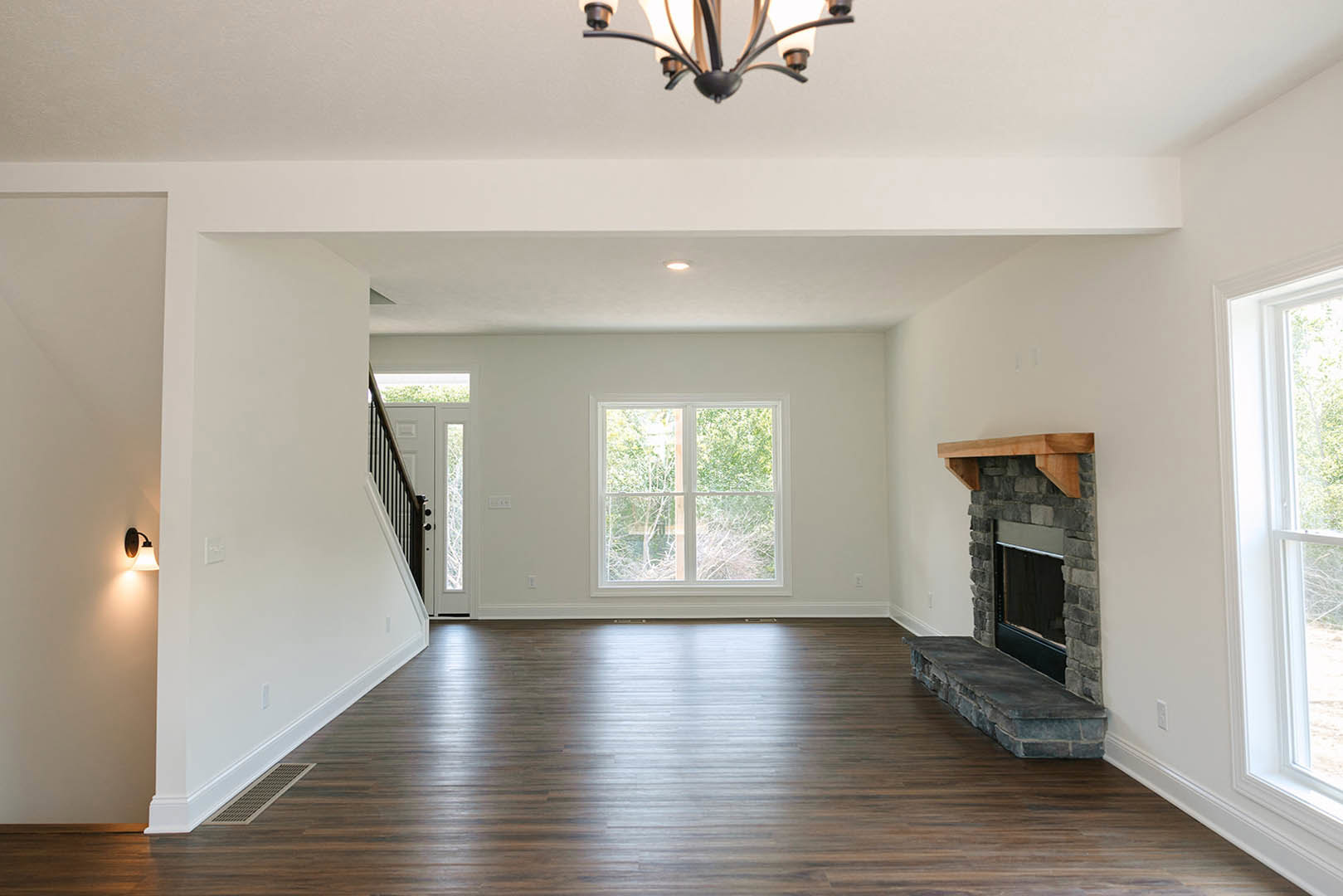 Living room with hardwood flooring, stone fireplace with ledge, large window showing trees outside, chandelier overhead, and stone bench along wood floor.