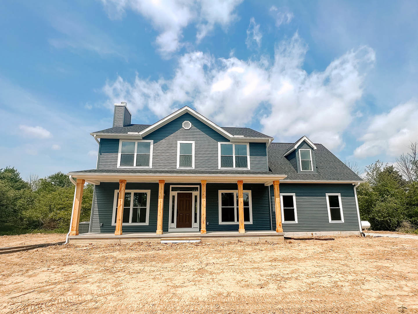 Partially built house with white-framed windows, brown glass-paneled door, covered front porch, exposed wooden posts, and dirt ground under clear blue sky with Grouseland visible
