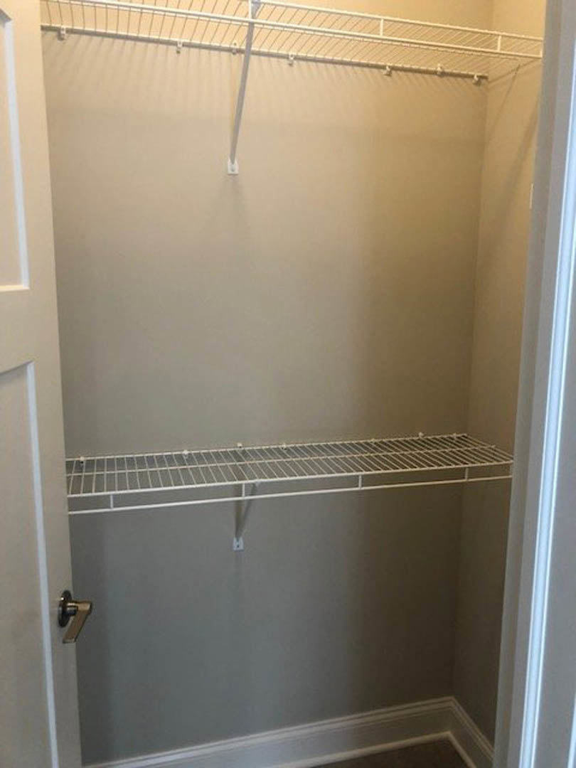 White metal shelf with horizontal bars mounted on plaster wall inside closet, adjacent to white door frame, with black object partially visible in foreground.