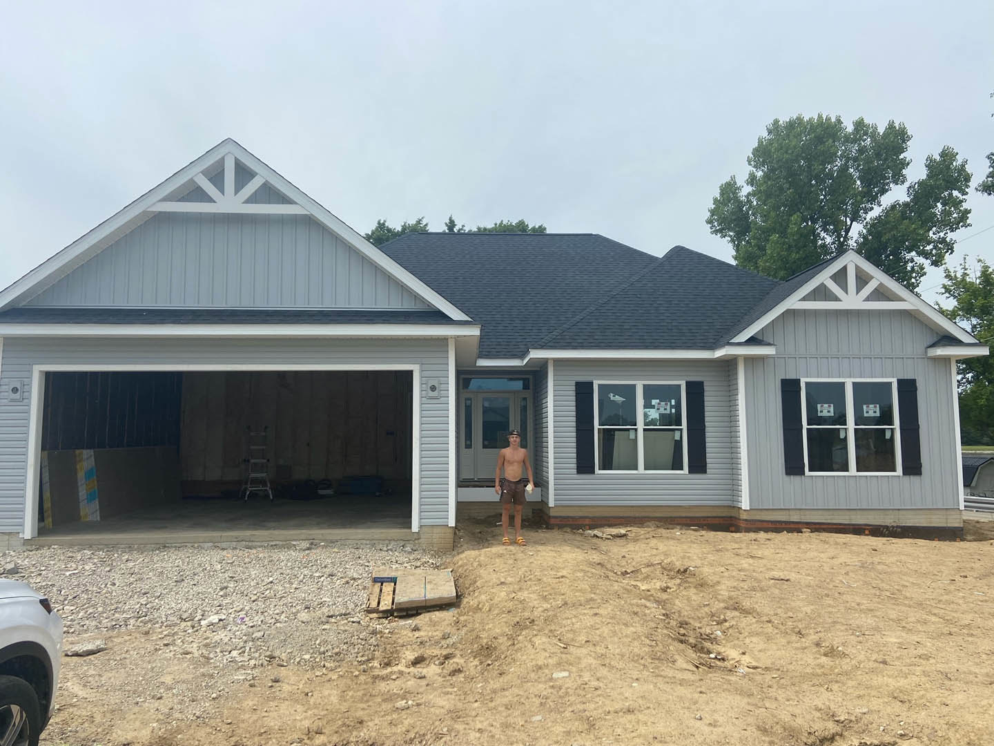 Man in shorts holding a bottle standing on driveway in front of modern home with white-framed windows, visible car tire, wall-mounted sign, and stickers on window