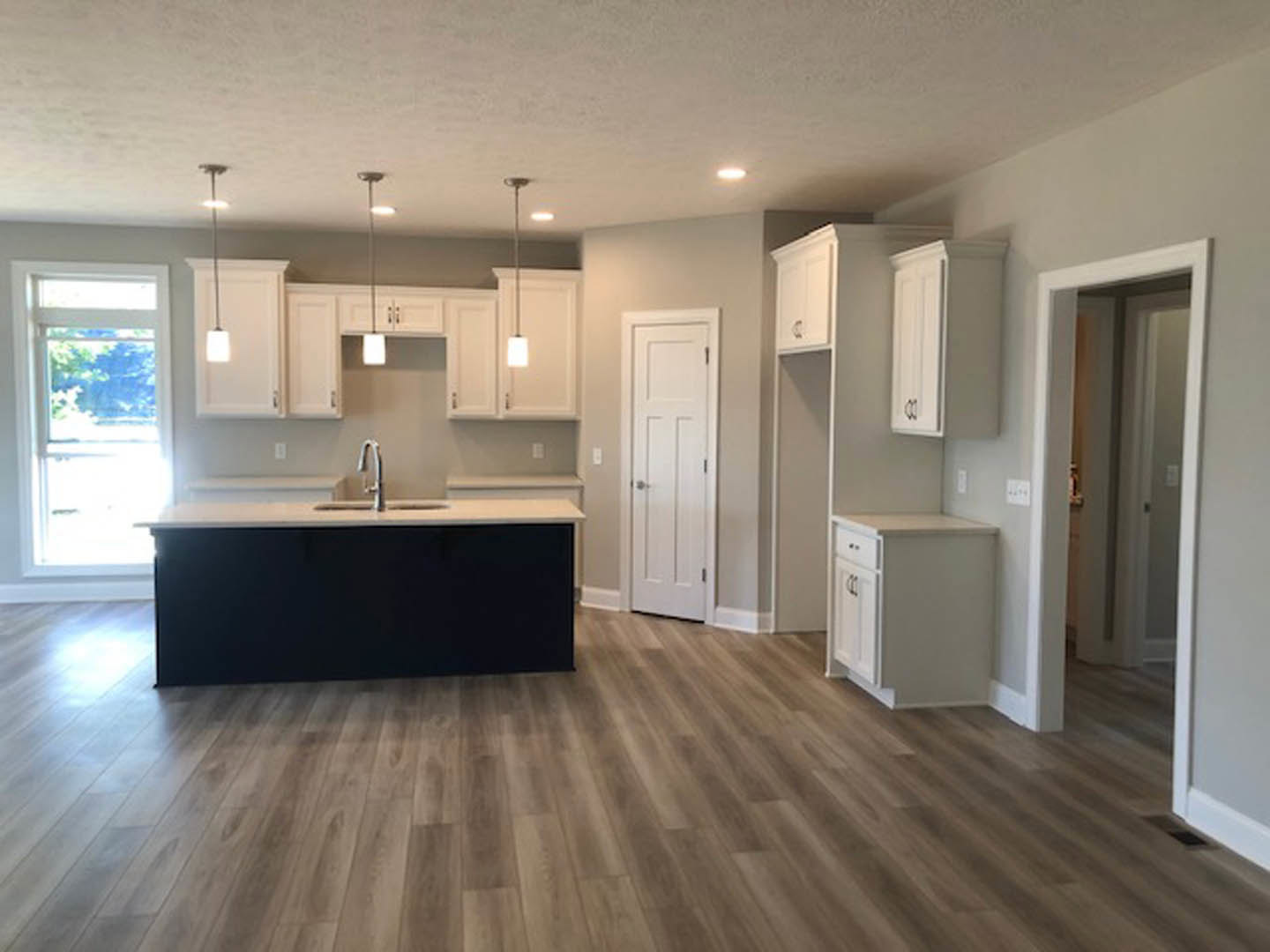 White kitchen cabinets with black countertops, silver faucet, wood flooring, white door with silver handle, large window letting in bright natural light.