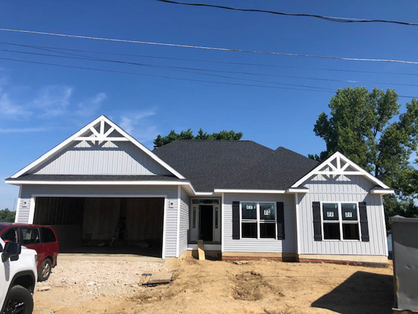 Partially built house with exposed framing, attached garage, red car parked in driveway, white SUV nearby, blue sign on white door, construction materials and waste container on
